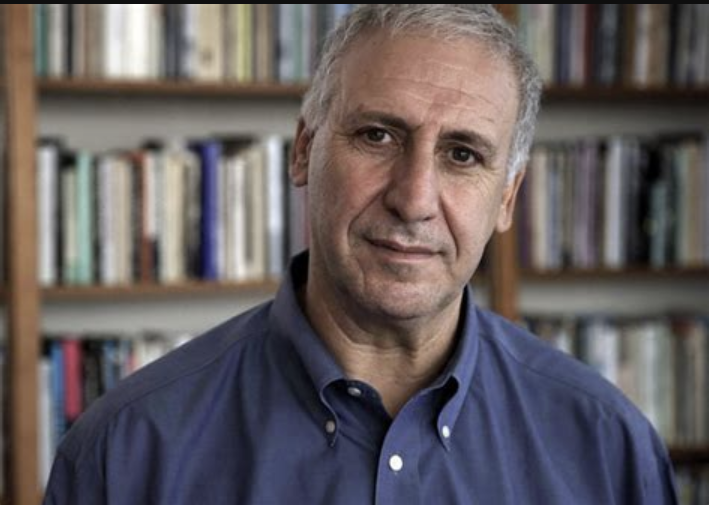 Edward Hirsch standing in front of bookcase wearing blue shirt and tie